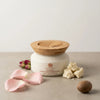 White jar with wooden lid on a beige surface with pink petals and a nutmeg.